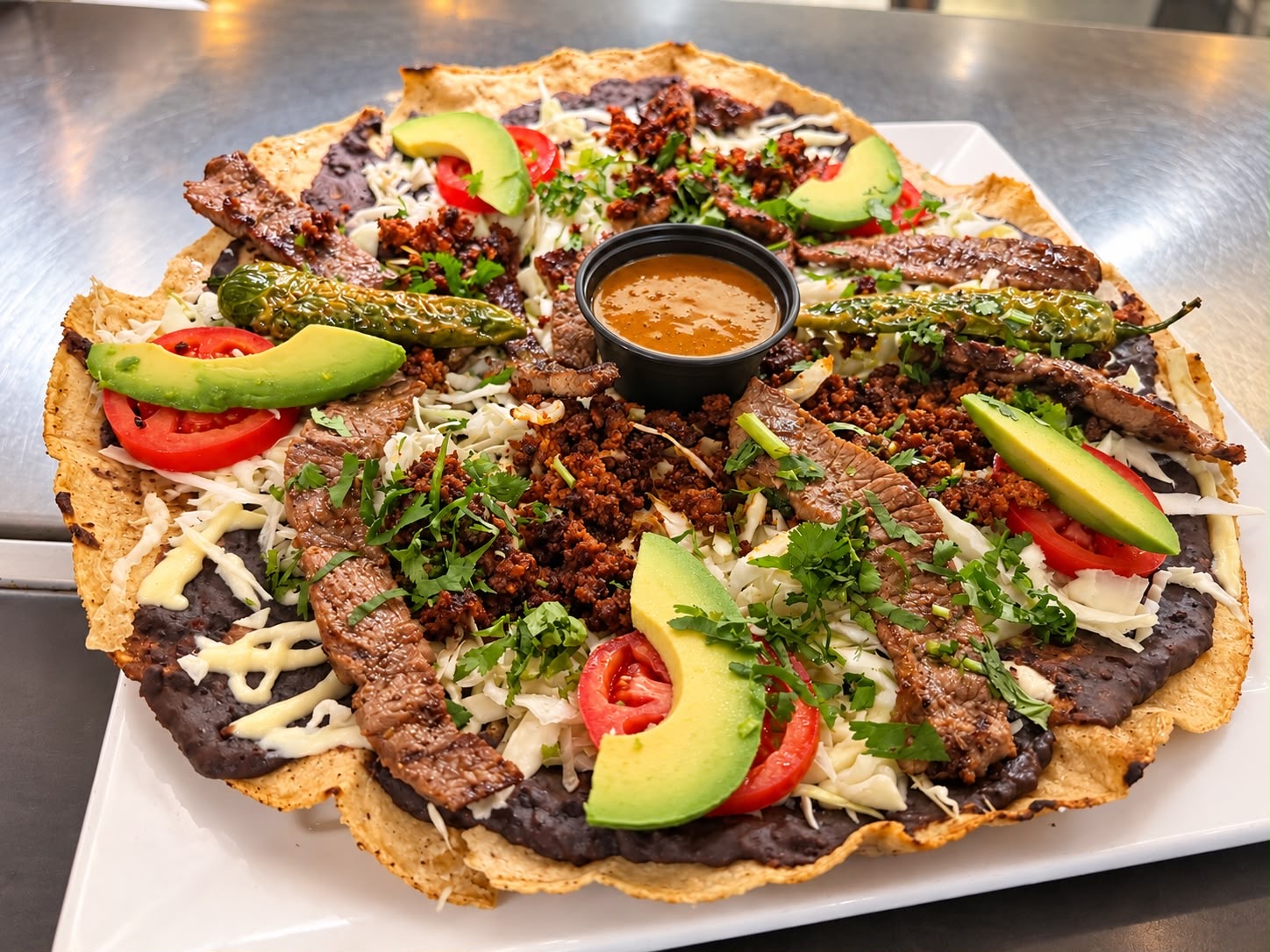 Giant crispy tostada layered with black beans, carne asada, chorizo, melted cheese, avocado, tomato and salsa