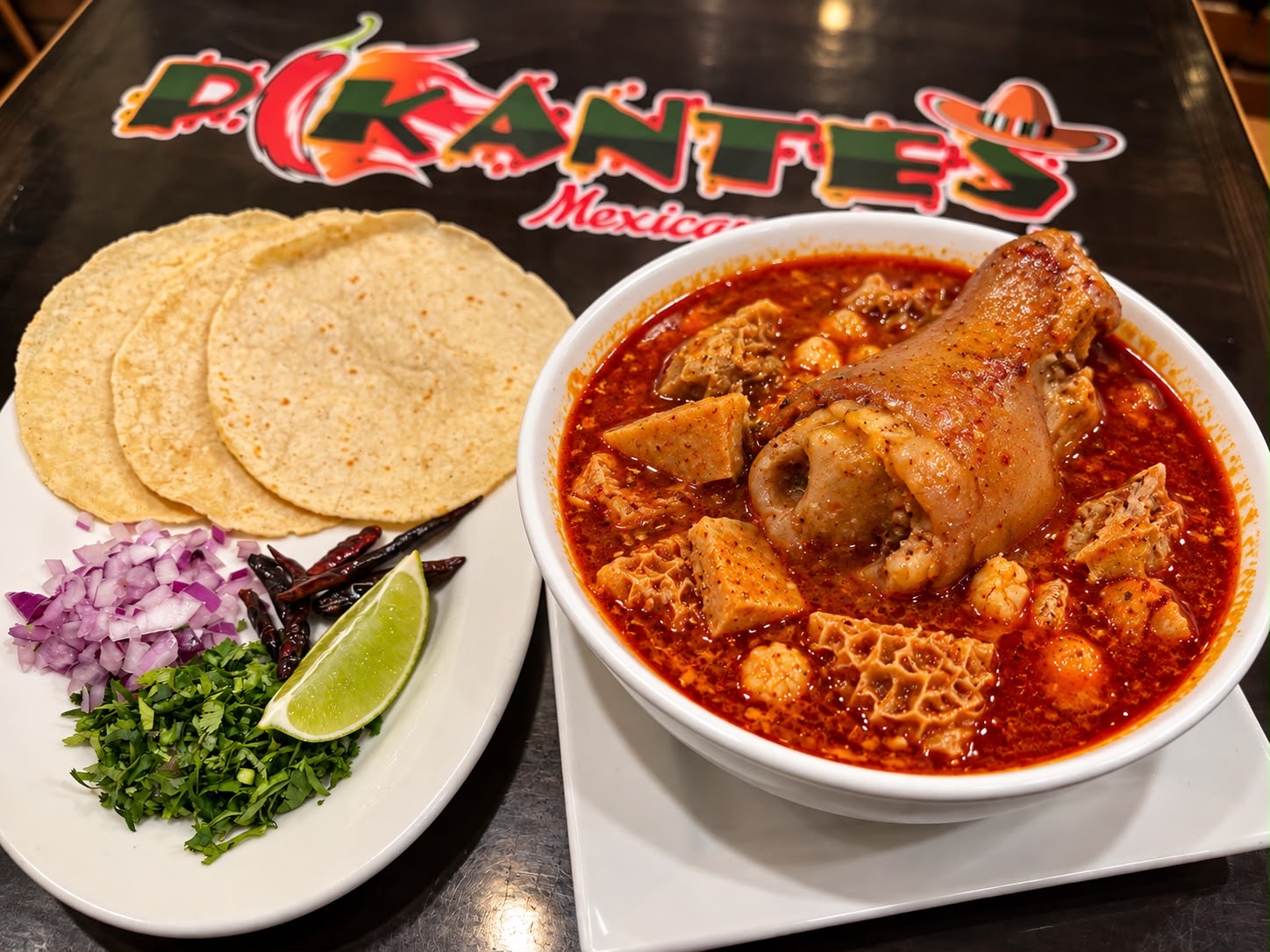 Bowl of red menudo with hominy and pig foot served with tortillas, onion, cilantro, chiles and lime