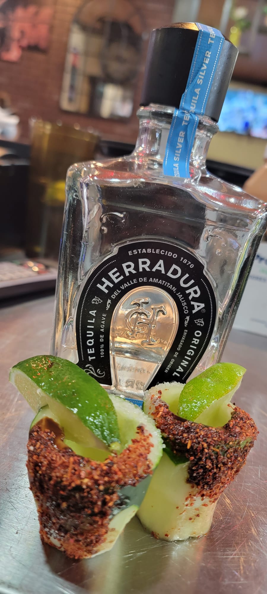 Two cucumber tequila shots with thick chili-salt rims and lime wedges beside a bottle of Herradura silver